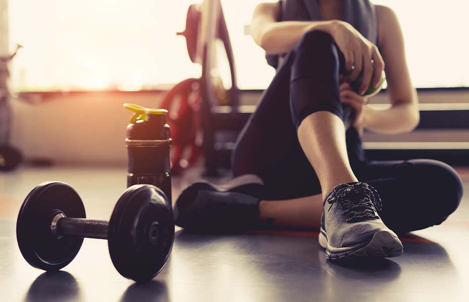 woman sitting on a gym floor next to a dumbbell and water bottle preparing to exercise.