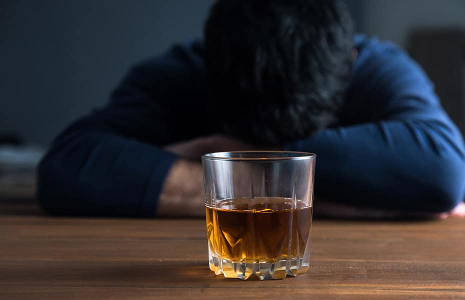 Man with his head down on a table in front of a glass of dark liquor.