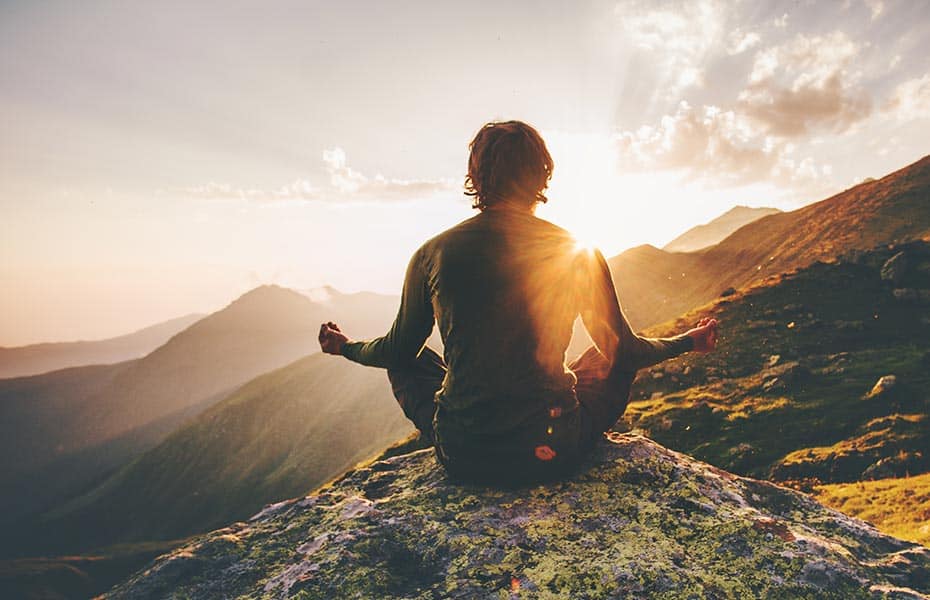 Man sitting cross-legged in green shirt meditating on mountain cliff overlooking the mountain side at sunrise. Reflects using meditation for spirituality.