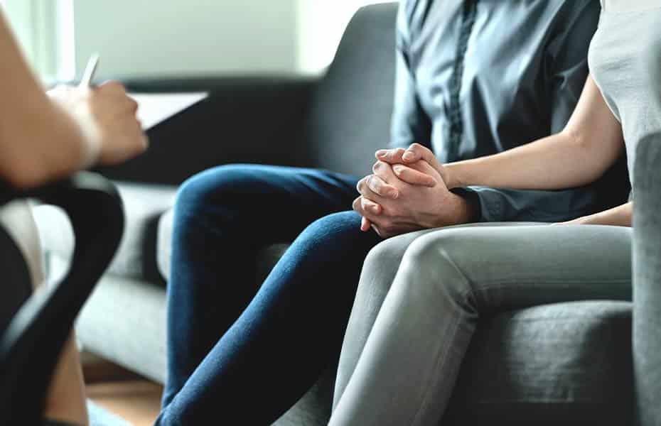 Man and woman holding hands while sitting with a therapist during couples rehab therapy session.
