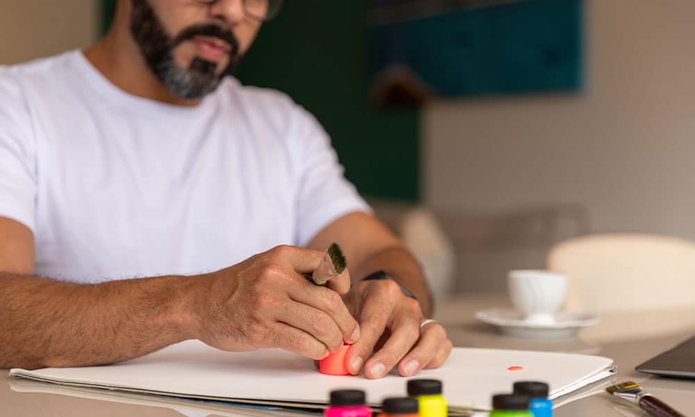 Bearded man sitting at table painting with acrylic paints during art therapy for addiction.