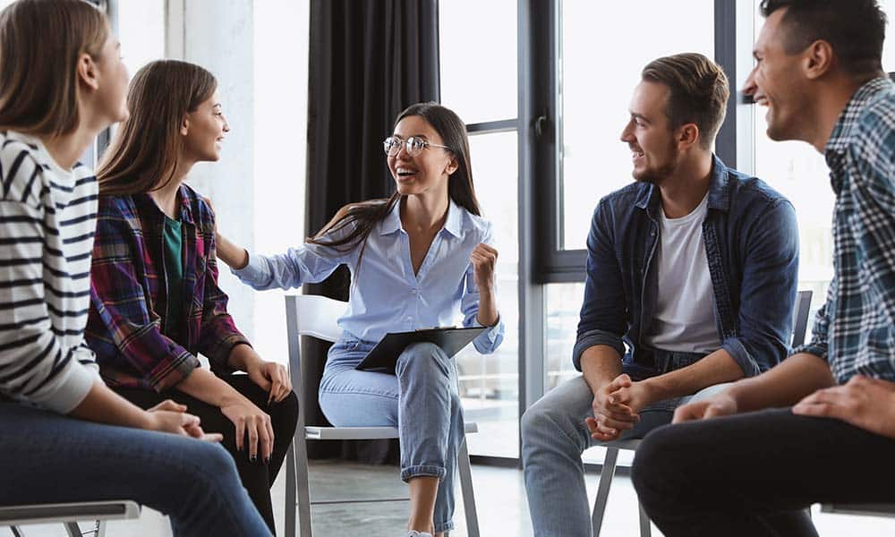 Happy coed group of addiction treatment patient sitting in a semi circle with a therapist who is practicing cognitive behavioral therapy with them.