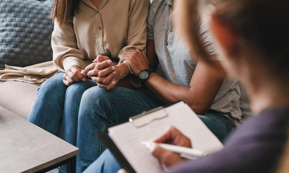 A couple sitting together holding hands while talking to their therapist during couples therapy in addiction treatment.