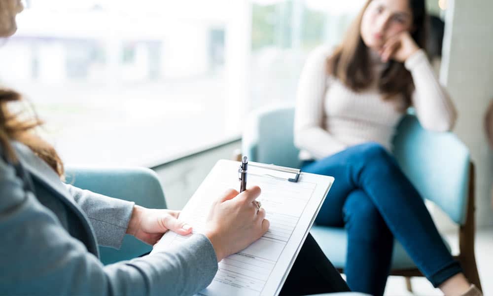 Female patient sitting in her therapists office during dialectical behavioral therapy session while her therapist writes on a notepad.