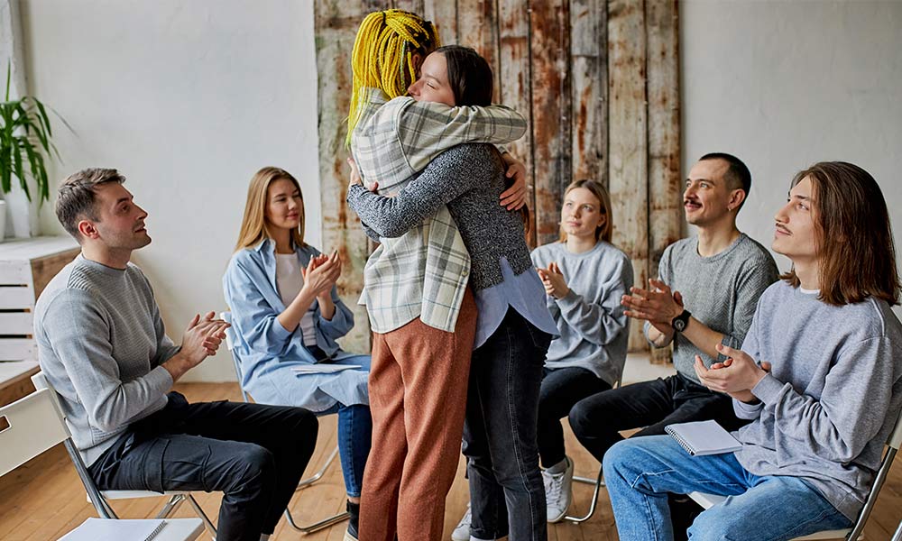 Coed group therapy session during addiction treatment. Five people sitting in a semi circle surrounding two of their group members who are giving each other a supportive hug.
