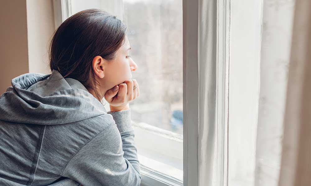 Woman alone staring out her window as she struggles with addiction and withdraws from her social circle.