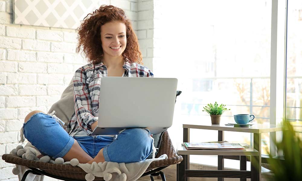 Woman with curly red hair sits in her living room chair with her laptop smiling as she attends online addiction treatment.
