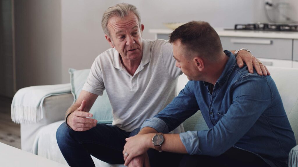 Father comforting his son on a couch while discussing the cost of rehab and insurance coverage options in Massachusetts.
